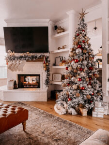 BURGUNDY AND GOLD CHRISTMAS DECORATING IDEAS | Living room decorated for Christmas with a Flocked Christmas tree, burgundy, gold, and silver ornaments, and a fireplace mantle decorated with flocked garland, bottle brush trees, large gold bells, and cream stockings.