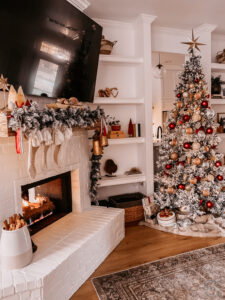 BURGUNDY AND GOLD CHRISTMAS DECORATING IDEAS | Living room decorated for Christmas with a Flocked Christmas tree, burgundy, gold, and silver ornaments, and a fireplace mantle decorated with flocked garland, bottle brush trees, large gold bells, and cream stockings.