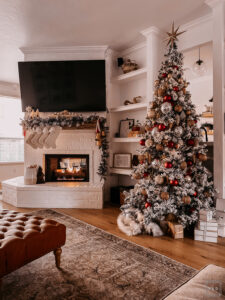 BURGUNDY AND GOLD CHRISTMAS DECORATING IDEAS | Living room decorated for Christmas with a Flocked Christmas tree, burgundy, gold, and silver ornaments, and a fireplace mantle decorated with flocked garland, bottle brush trees, large gold bells, and cream stockings.